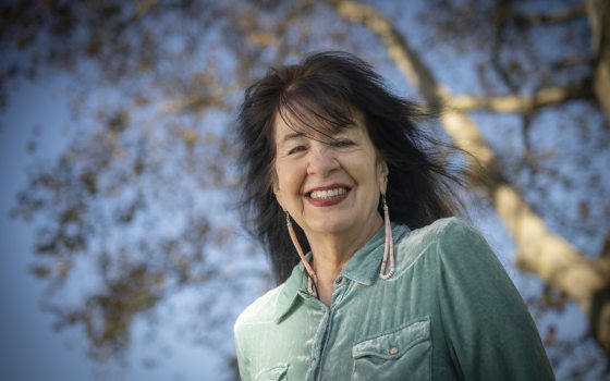 Outdoor portrait of poet Joy Harjo smiling at the camera, wearing a light teal velvet collared shirt and long beaded earrings, with a large tree and clear blue sky out of focus behind her.